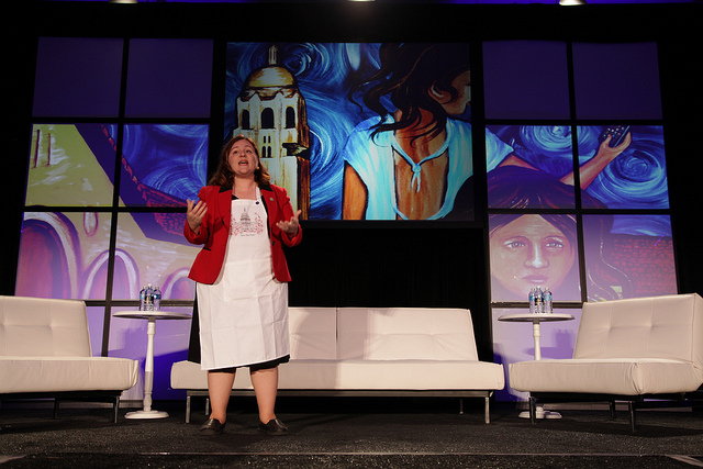 A woman wearing an artist's apron stands on stage in front of images of her paintings