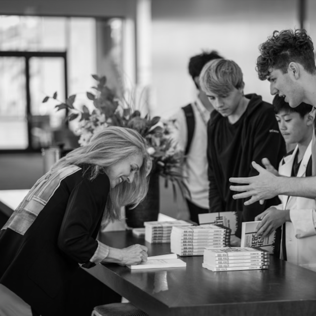 A woman leans over a table as she signs a book. Young people gather on the other side of the table, which has books stacked on it.