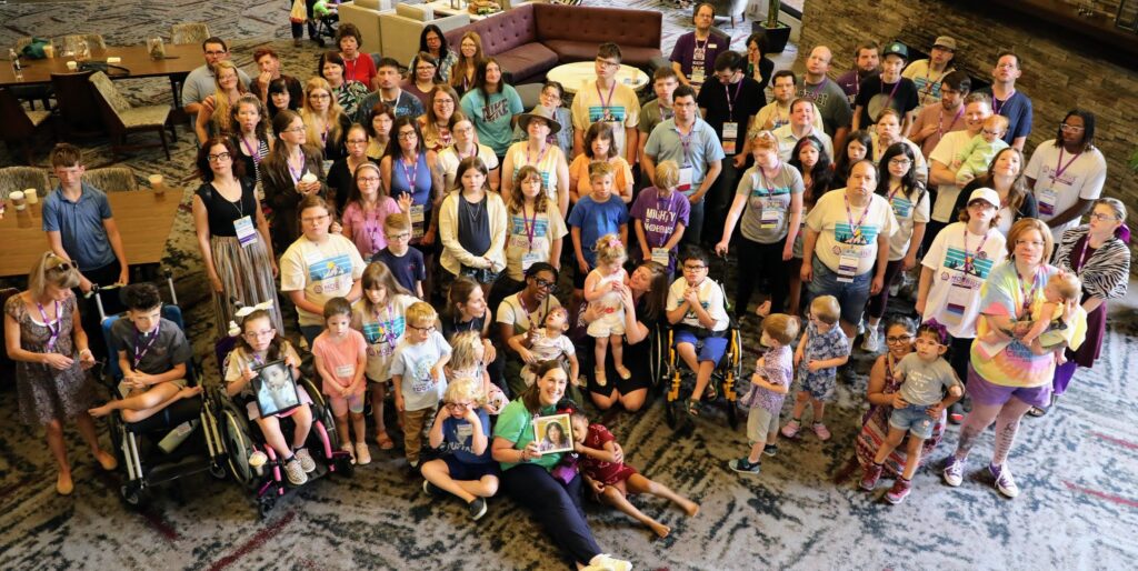 About 60 adults and children look up at the camera. A few are in wheelchairs and one person in the front row is holding a photo of a person.